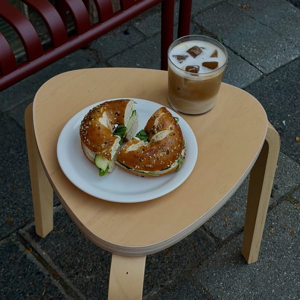 Belegter Bagel mit Gurke und Salat auf einem Teller, daneben ein Eiskaffee, auf einem Holzhocker im Freien