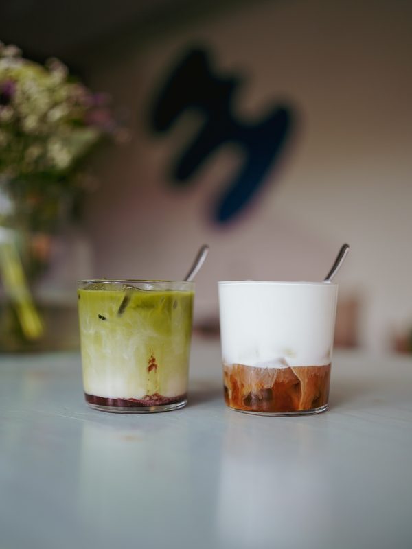Zwei Gläser auf einem Tisch im mela: ein grüner Matcha mit Fruchtschicht und ein Eiskaffee mit Milchschaum, dahinter unscharf das mela-m an der Wand